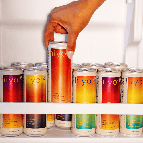 Hand holding a tall Hiyo beverage can surrounded by various colorful canned drinks in red, yellow, and orange flavors on whit