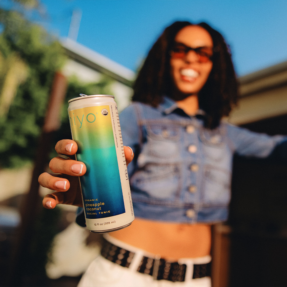 Tall slim can with gradient color transitioning from yellow to teal, labeled "pineapple coconut facial tonic," held in hand o