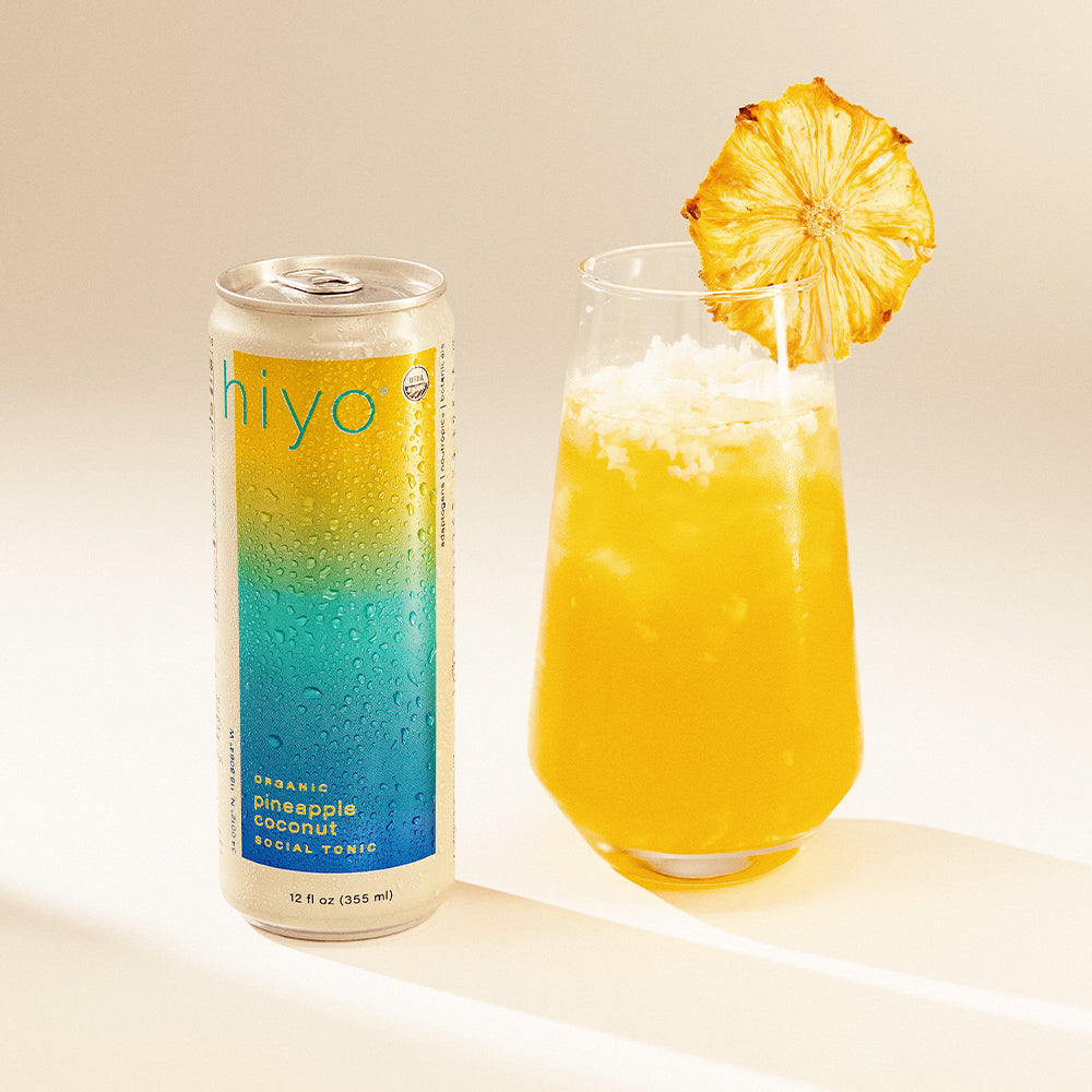 Closed aluminum can and poured glass of yellow beverage garnished with pineapple slice on tan background.