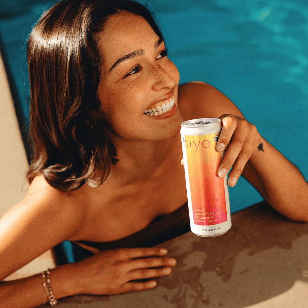 Woman holding tall clear can with gradient yellow-to-red liquid labeled "iivo" by poolside.