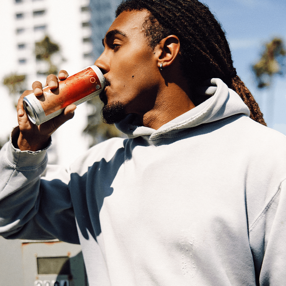 Man in white hoodie drinking strawberry guava beverage from can outdoors.