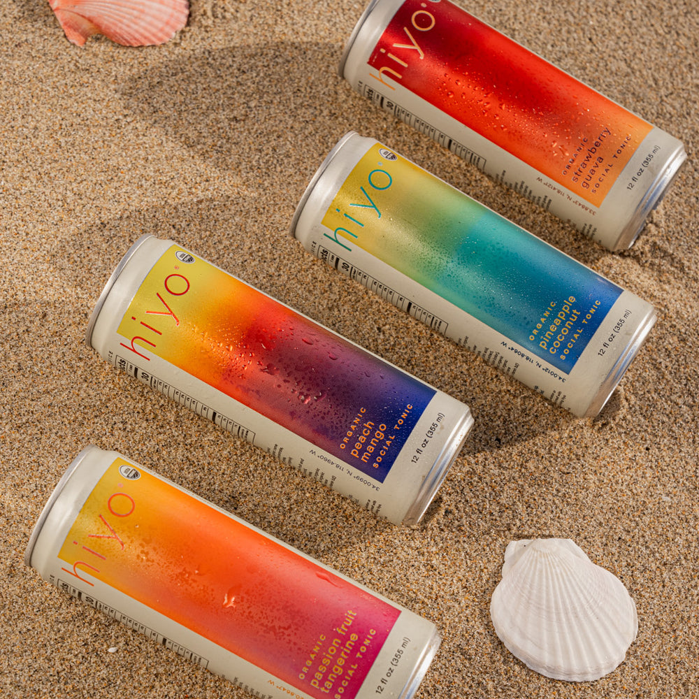 Four colorful beverage cans with gradient designs arranged on sand with a white seashell.