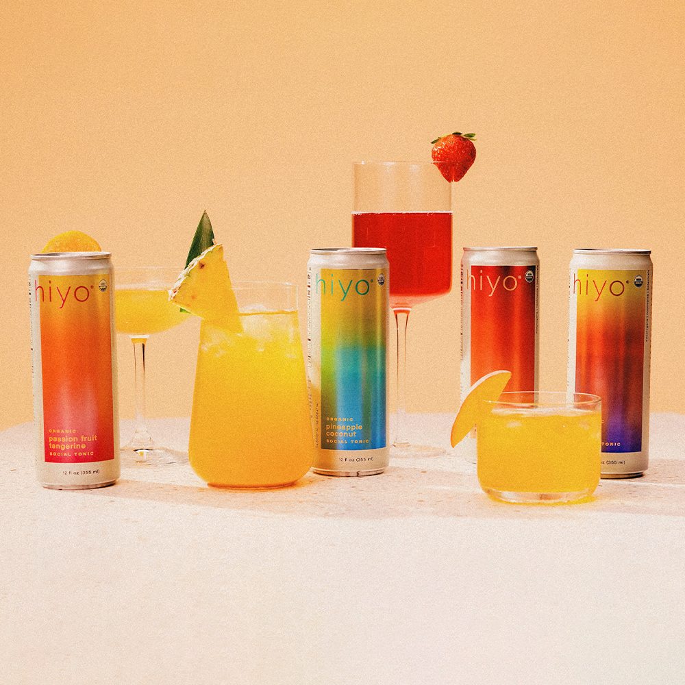 Five colorful Iyo beverage cans and glasses with tropical juices, citrus slices, and a strawberry garnish on beige background