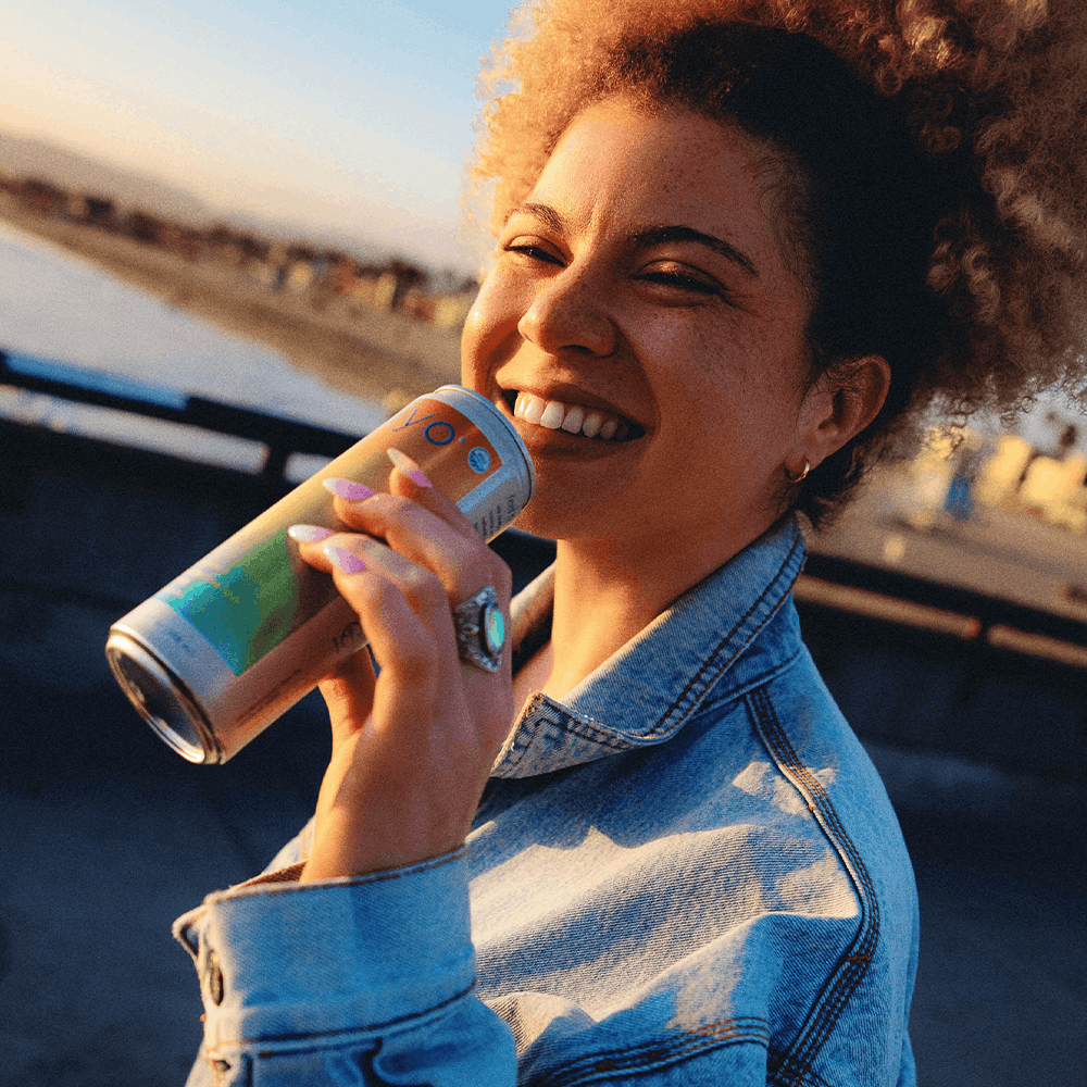 Person holding and drinking from a blue and green labeled beverage can outdoors.