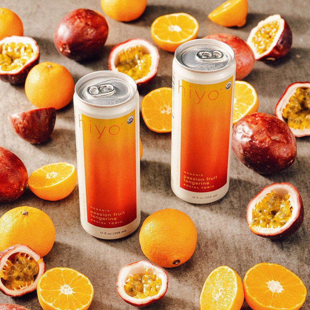 Two cans of non-alcoholic functional seltzer hiyo passion fruit tangerine on a surface with tangerines and passion fruits.