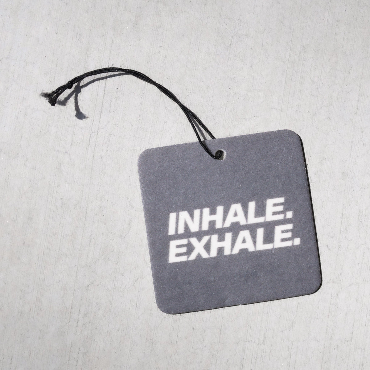 Gray square air freshener with "INHALE. EXHALE." text and black string attachment.
