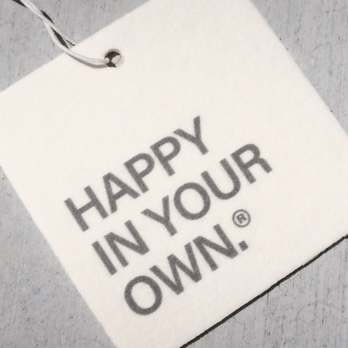 White rectangular air freshener tag with gray text reading "Happy in your own" and a registered trademark symbol, attached wi