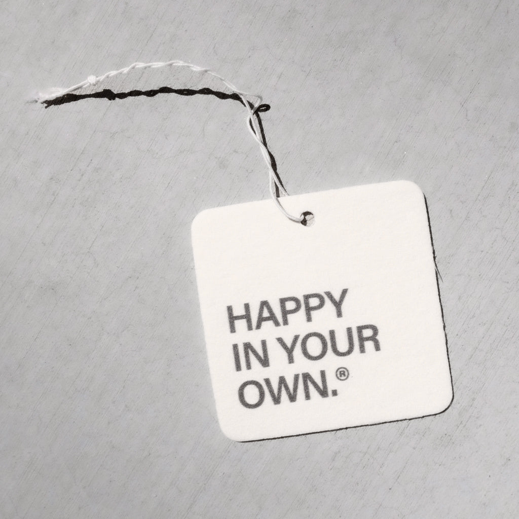 Square white air freshener tag with "HAPPY IN YOUR OWN" text, attached with black and white twine.