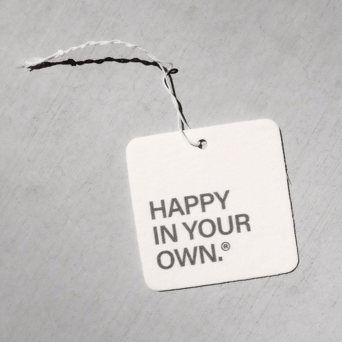 Square white air freshener tag with "HAPPY IN YOUR OWN" text, attached with black and white twine.