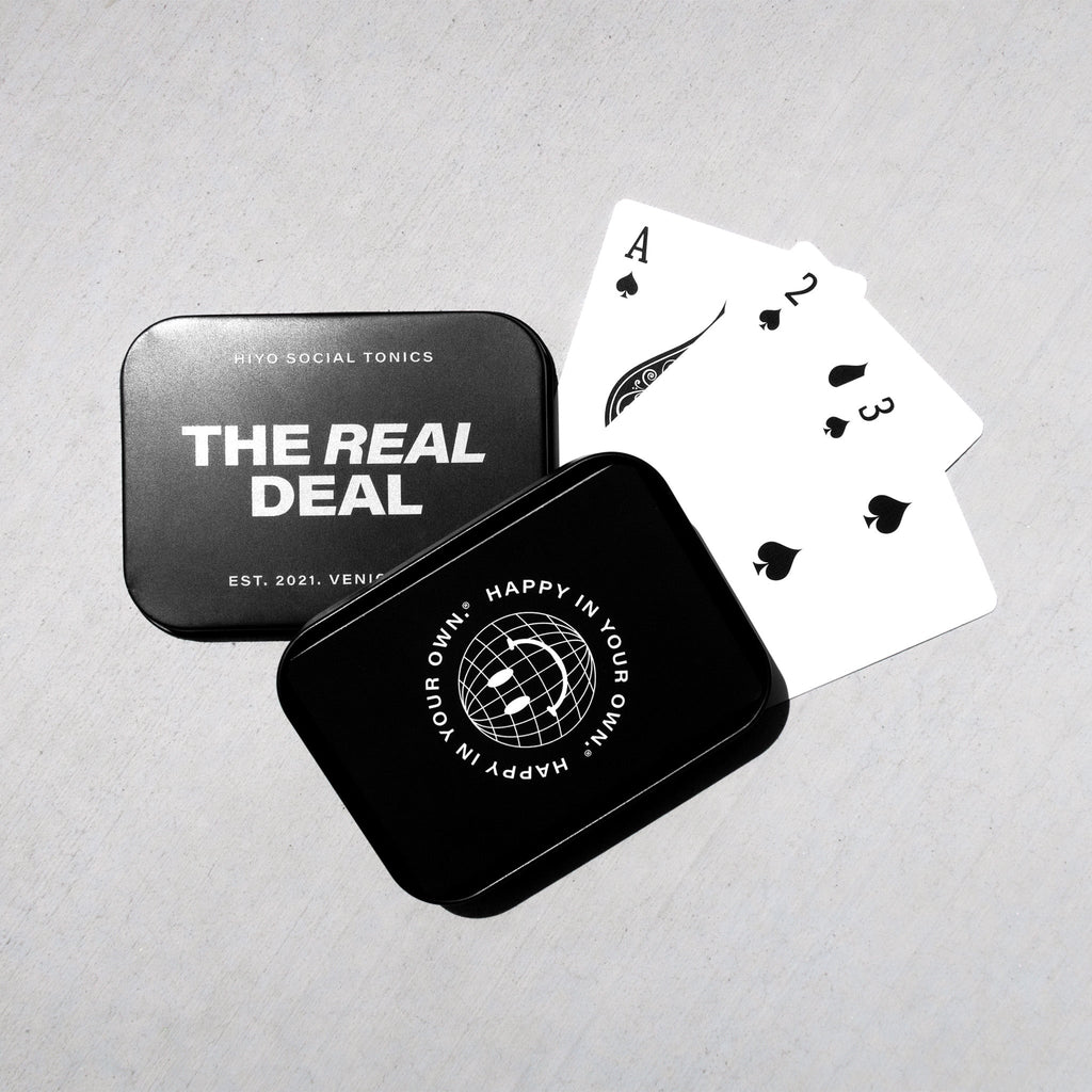 Black and white playing cards with spade suit displayed next to two metal storage tins labeled "The Real Deal" and "Happy in 