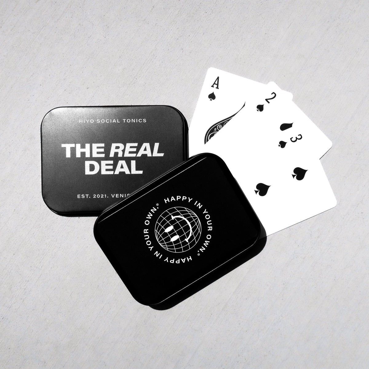 Black and white playing cards with spade suit displayed next to two metal storage tins labeled "The Real Deal" and "Happy in 
