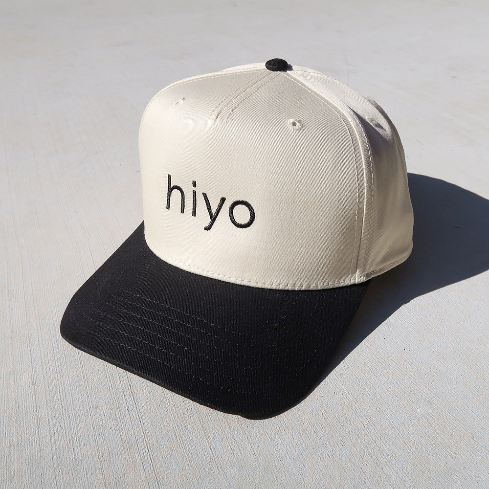 Cream and black trucker hat with "hiyo" text embroidered on the front and mesh panels on the sides.
