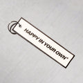 Rectangular white keychain with black text reading "HAPPY IN YOUR OWN" and metal ring attachment on light textured background