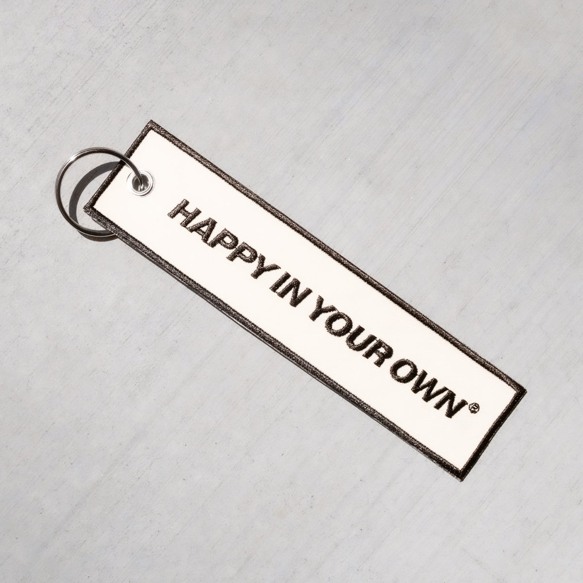 Rectangular white keychain with black text reading "HAPPY IN YOUR OWN" and metal ring attachment on light textured background