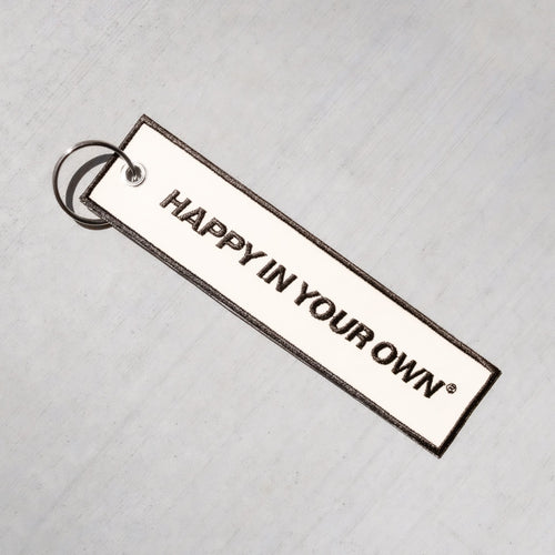 Rectangular white keychain with black text reading "HAPPY IN YOUR OWN" and metal ring attachment on light textured background