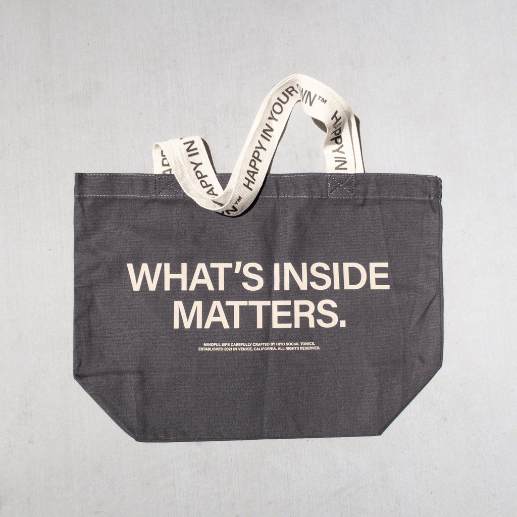 Charcoal tote bag with "WHAT'S INSIDE MATTERS" text and cream-colored handles with repeated text print.