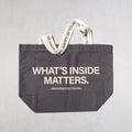 Charcoal tote bag with "WHAT'S INSIDE MATTERS" text and cream-colored handles with repeated text print.