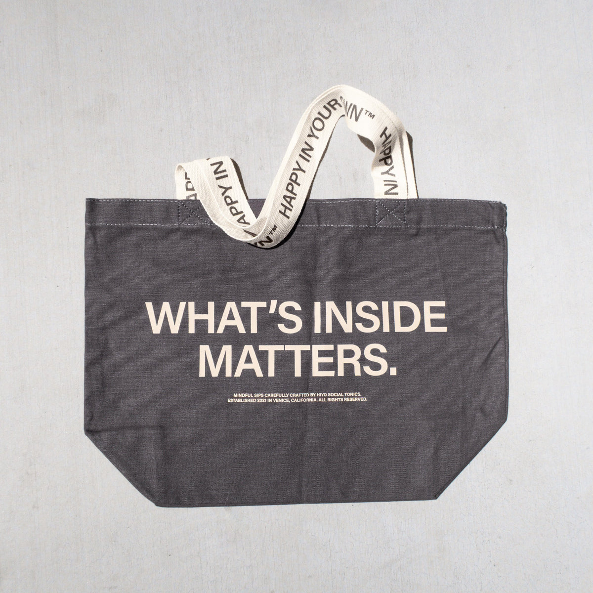 Charcoal tote bag with "WHAT'S INSIDE MATTERS" text and cream-colored handles with repeated text print.