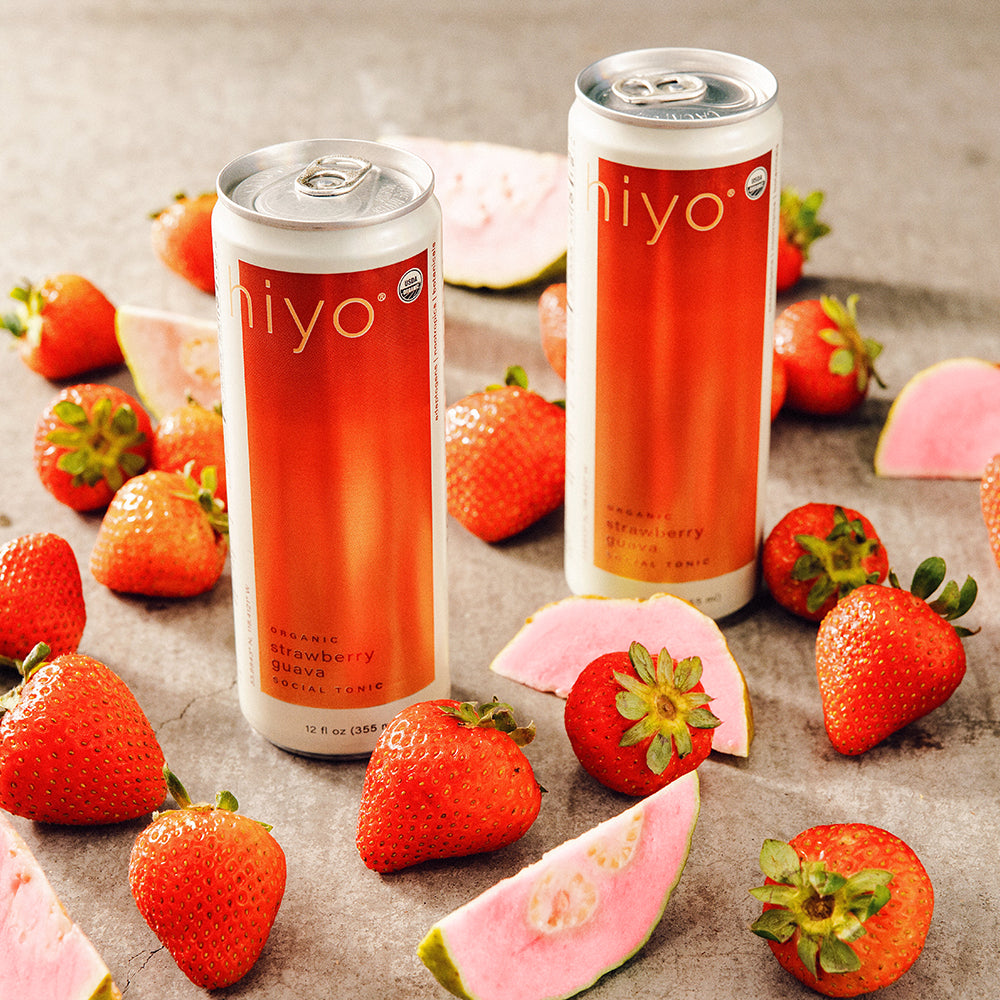 Two cans of non-alcoholic functional seltzer hiyo strawberry guava drink surrounded by strawberries and guava on a textured surface.