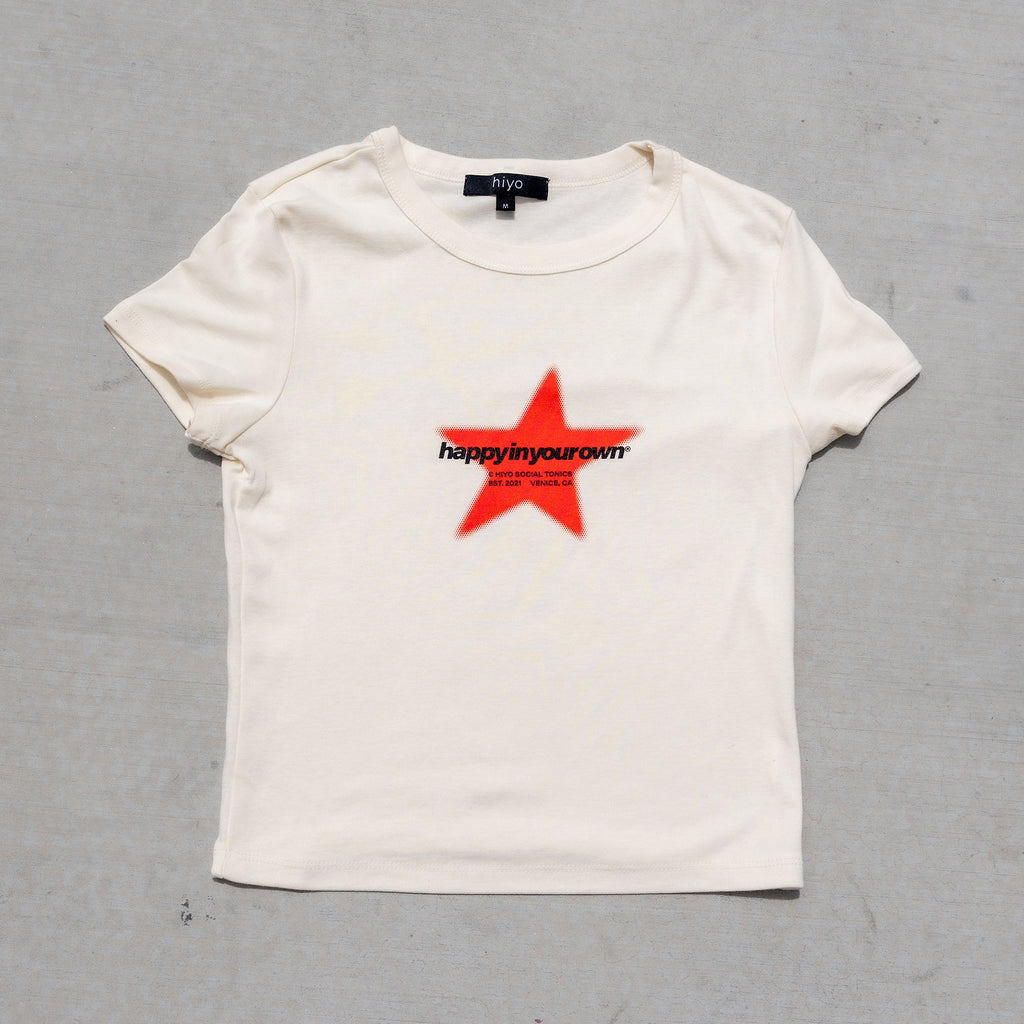 Cream baby tee with red star and "happy in your own" text graphic on chest.