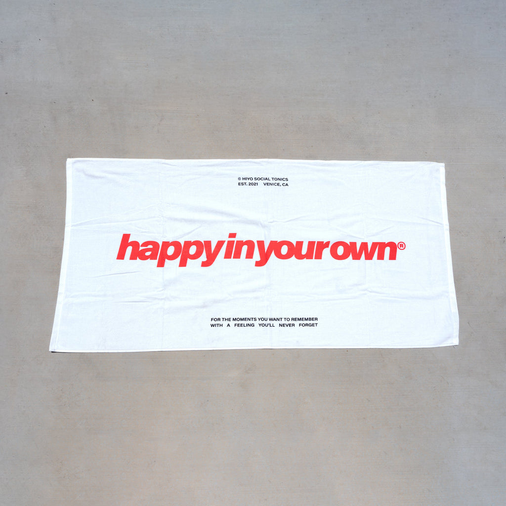 White beach towel with red "happyinyourown" text and tagline printed on front.