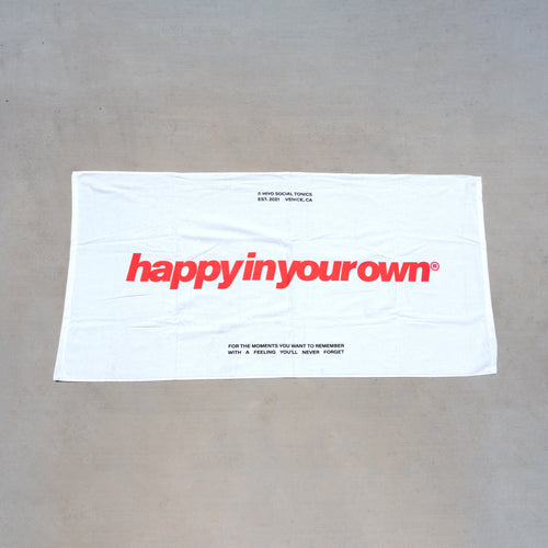White beach towel with red "happyinyourown" text and tagline printed on front.