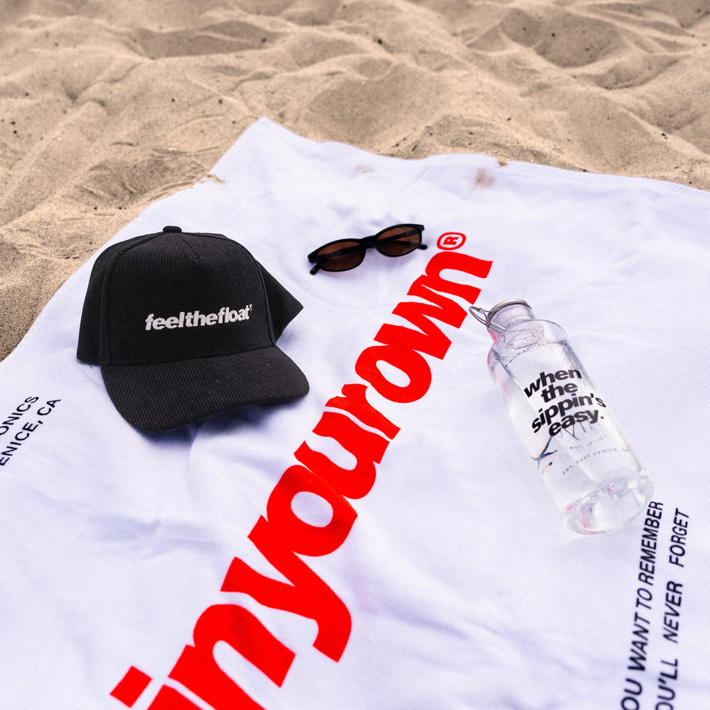 White beach towel with large red text displayed on sand, styled with black cap, sunglasses, and water bottle.