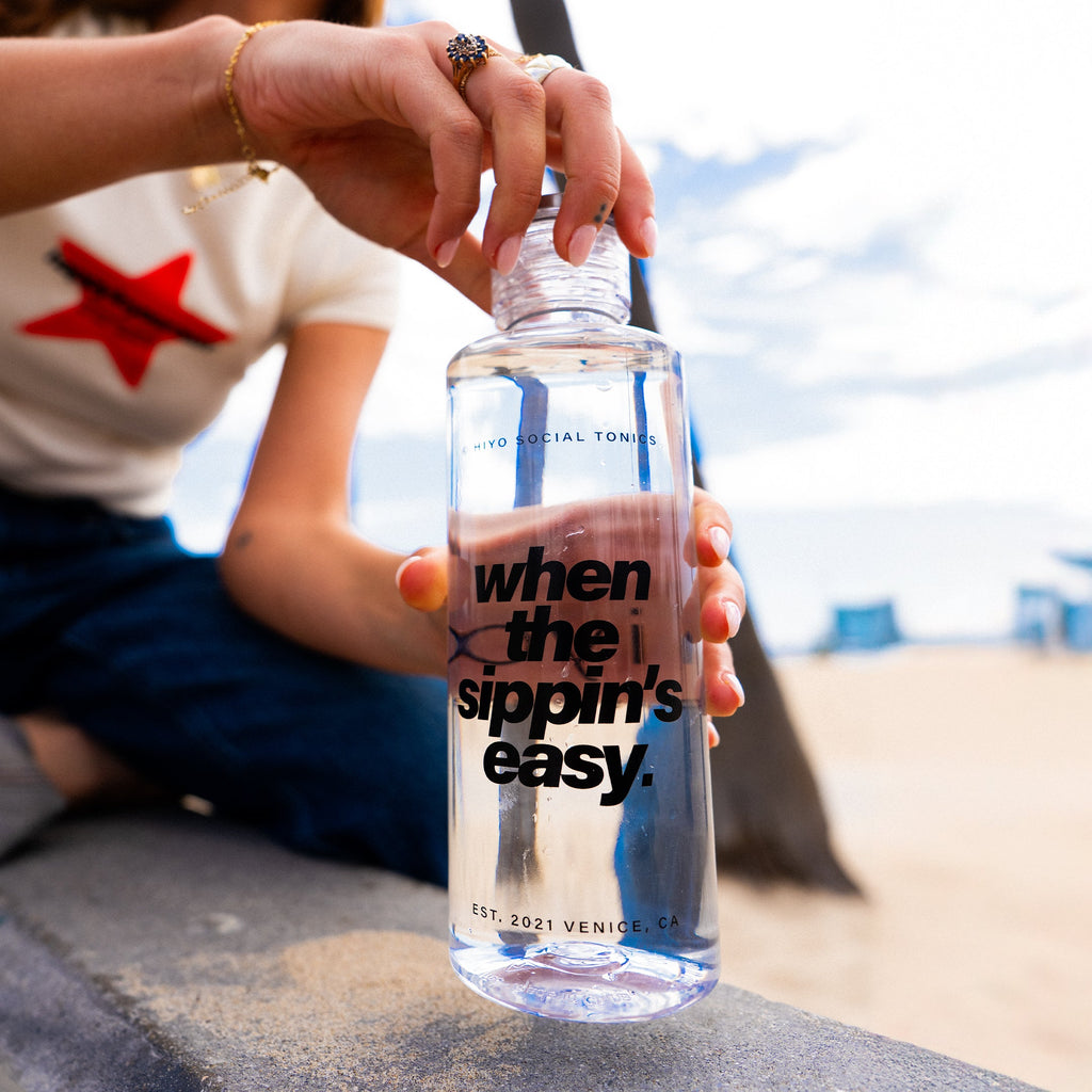 Clear water bottle with "when the sippin's easy" text held by person at beach with blue striped design.