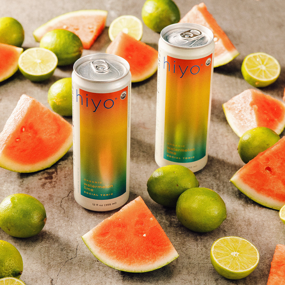 Two cans of non-alcoholic functional seltzer hiyo watermelon lime  surrounded by watermelon slices and limes on a textured surface.