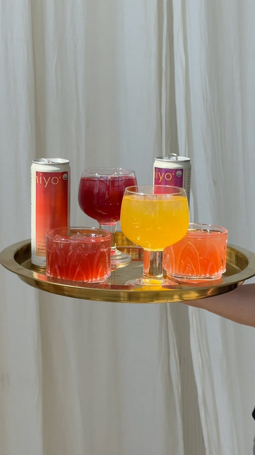 close-up of a hand opening a can of hiyo blackberry lemon social tonic in warm sunlight. the drink is poured into a stemmed glass over ice, creating a vibrant pink beverage with a foamy top. the video ends with the finished drink served on a gold tray alongside hiyo cans in blackberry lemon and other flavors.