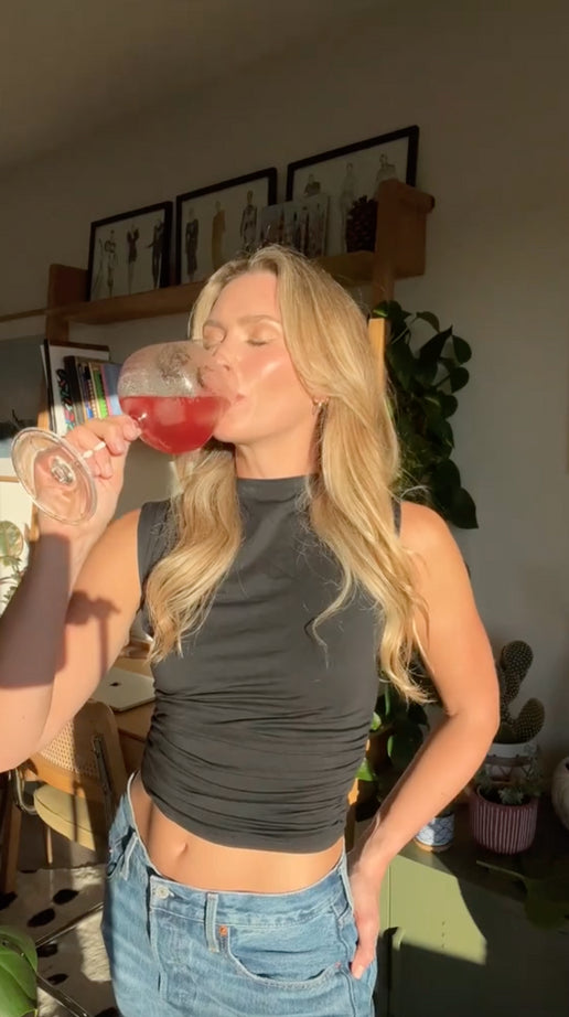 a video of a woman sitting on her living room floor, facing the camera and well lit by the sun with a can of strawberry guava hiyo in her hand. she has long blonde hair, hazel eyes, and is wearing a black top.